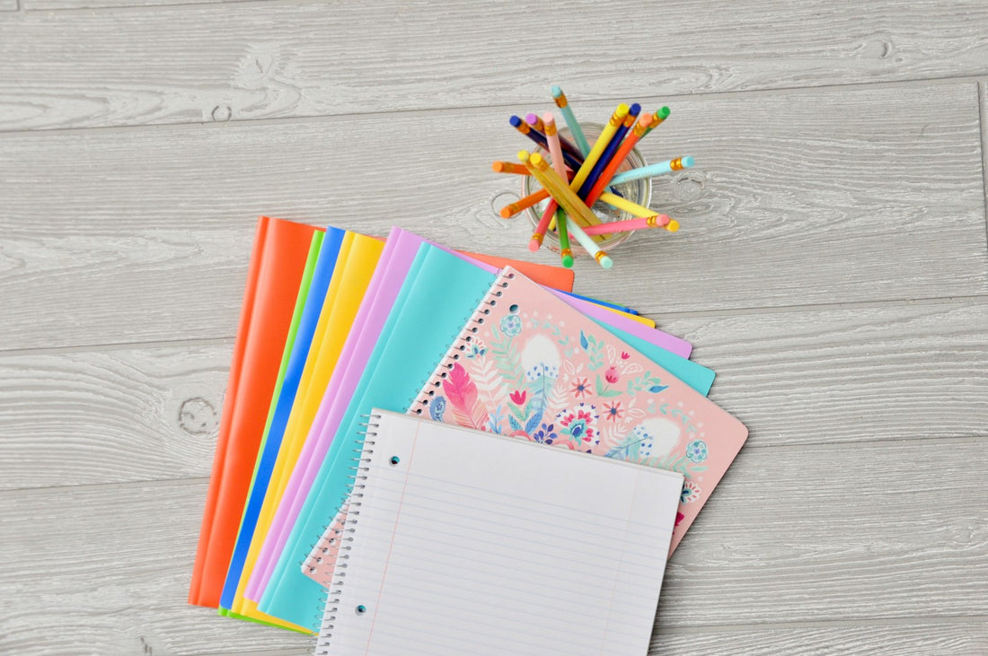 Fournitures scolaires en éventail sur un plan de bois clair : cahiers à spirale colorés, bloc ligné vierge et pot de crayons de couleur