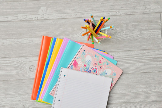 Fournitures scolaires en éventail sur un plan de bois clair : cahiers à spirale colorés, bloc ligné vierge et pot de crayons de couleur