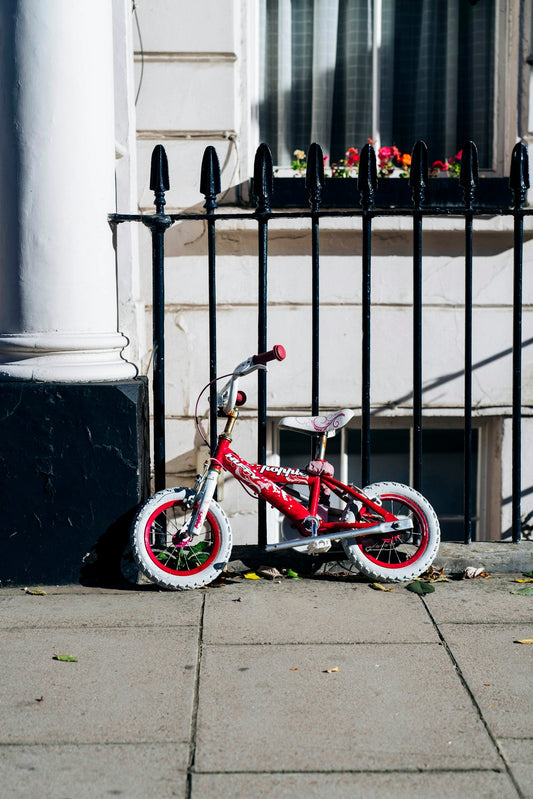 Petit vélo d'enfant rouge et blanc appuyé contre une grille, roues cerclées de rouge, prêt à recevoir des stickers d'enjoliveur