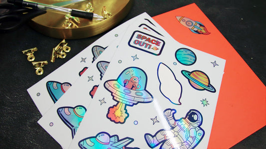 Planche d'autocollants holographiques sur le thème de l'espace posée sur une table d'atelier, avec ciseaux et pinces