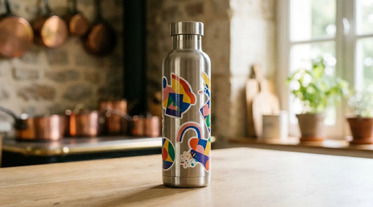 Gourde isotherme en inox brossé décorée d'autocollants aux motifs abstraits colorés, posée sur une table en bois dans une cuisine lumineuse