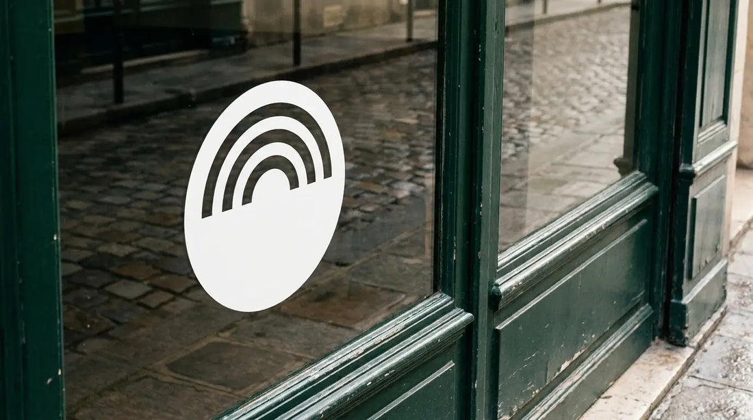 Sticker logo d'entreprise en adhésif blanc appliqué sur la vitrine d'un commerce à devanture verte, la rue pavée se reflétant dans le verre