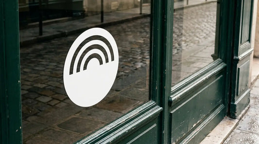 Sticker logo d'entreprise en adhésif blanc appliqué sur la vitrine d'un commerce à devanture verte, la rue pavée se reflétant dans le verre